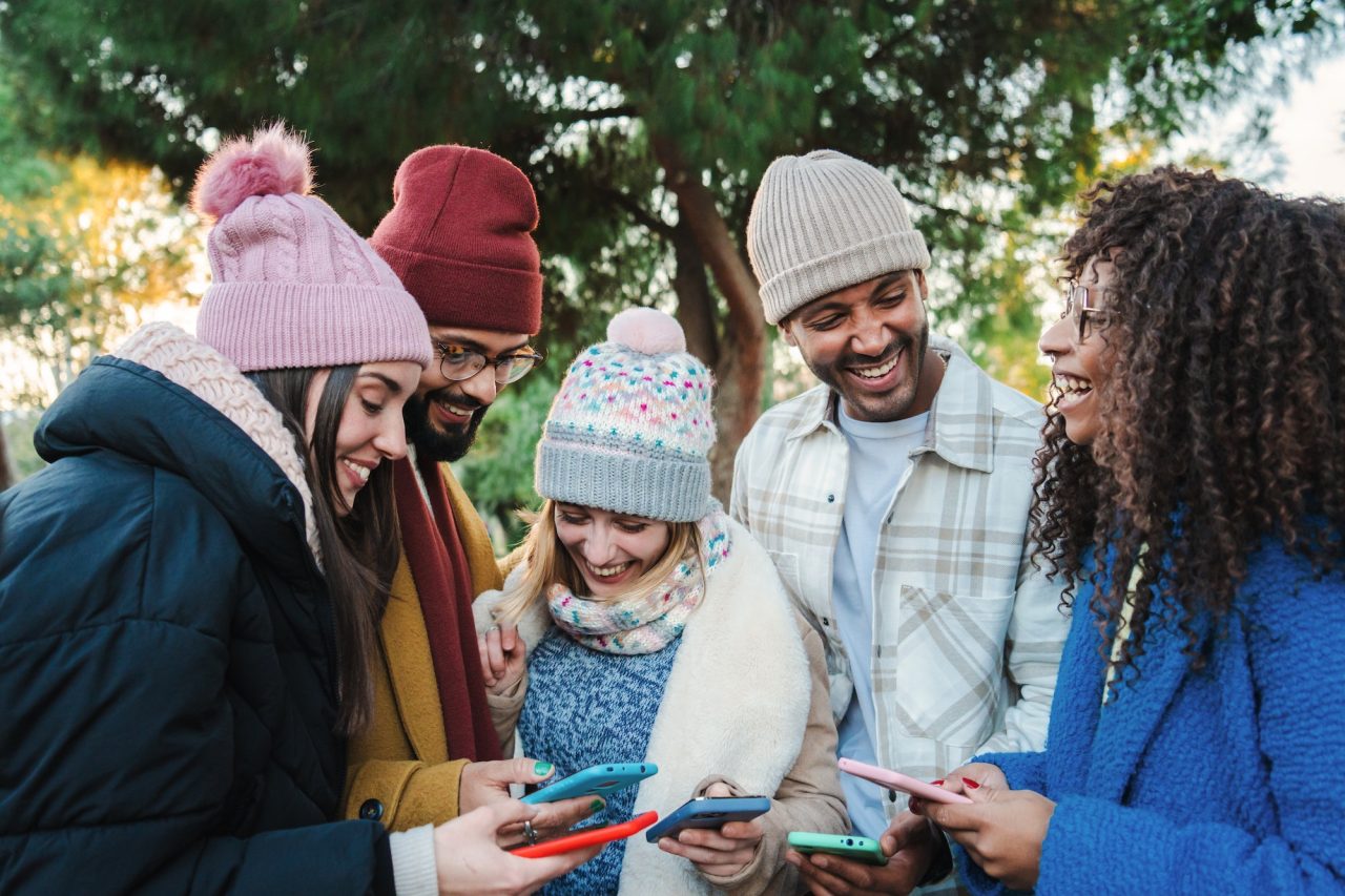 group of multiracial young friends with coats and hats smiling and watching the social media with a